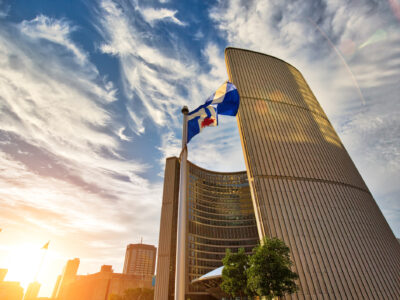 Photograph of a Toronto City flag blowing in the wind in front of city hall while the sun sets behind it.