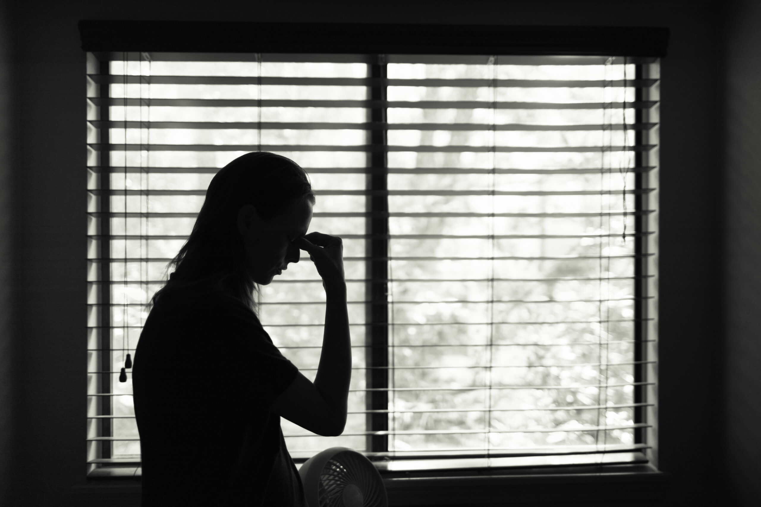 A silhouette of a stressed woman standing next to a bedroom window with her hand on her forehead.