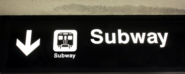 Photo of a black sign hanging from the ceiling of a subway tunnel with a white arrow pointing down and the words "Subway" written on it.