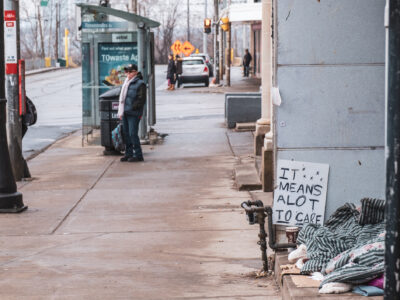 Scene of houseless person with a gratitude sign next to makeshift campsite along a sidewalk in Parkdale. A pedestrian waits for a streetcar in background.