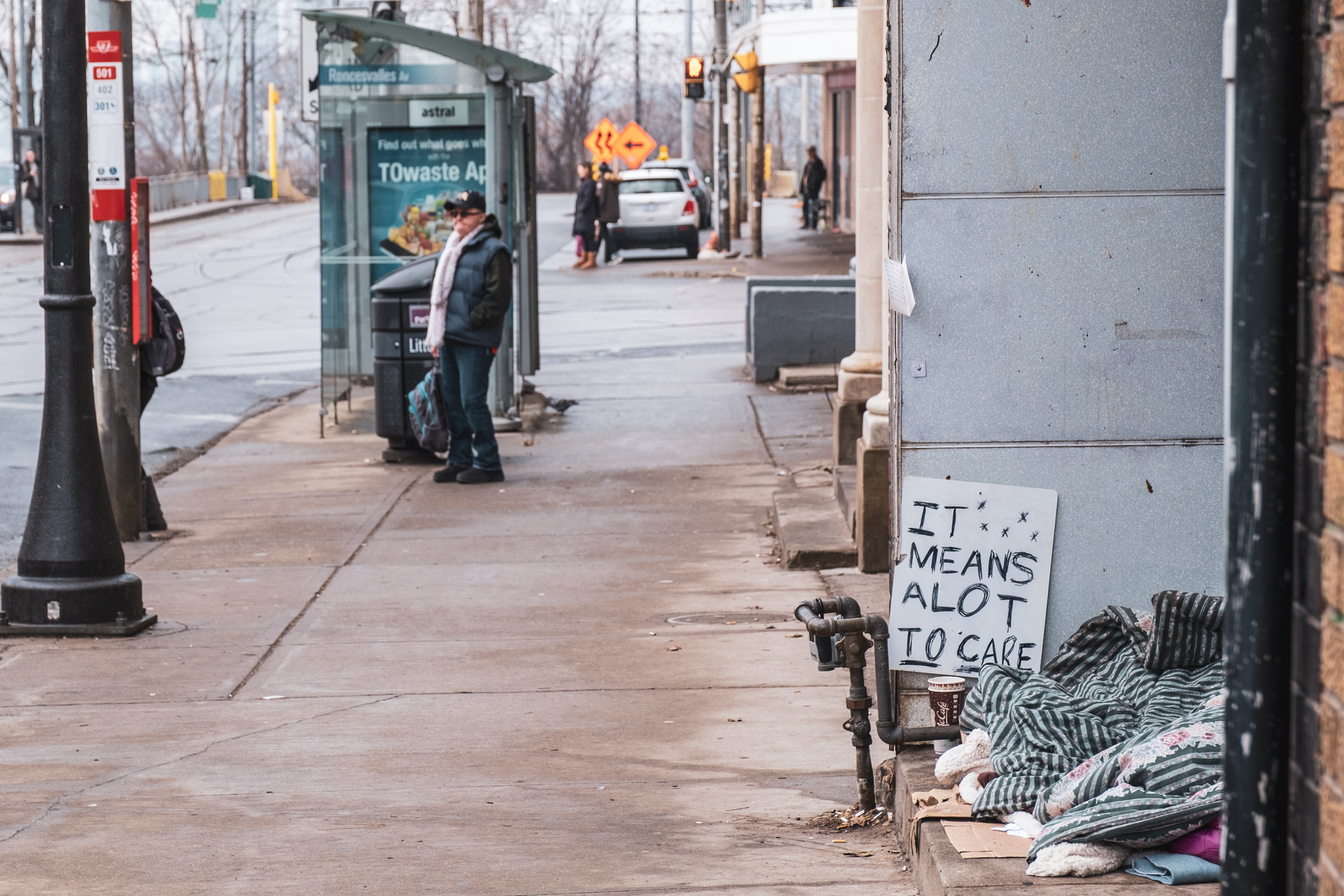 Scene of houseless person with a gratitude sign next to makeshift campsite along a sidewalk in Parkdale. A pedestrian waits for a streetcar in background.