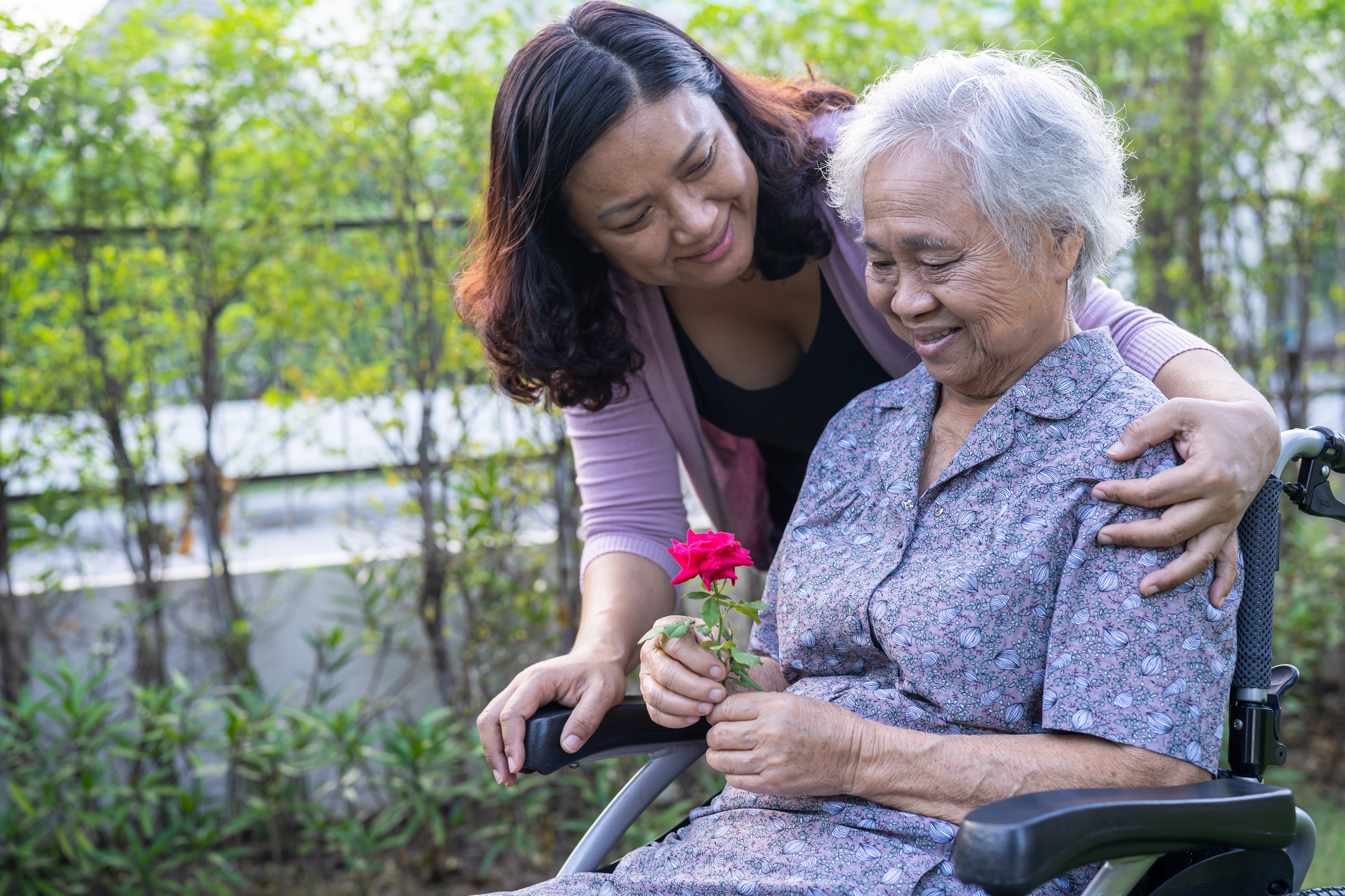 An Asian woman wraps her arm around her mother's shoulders, who is sitting in a chair smiling while holding a red flower.