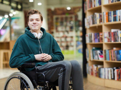 Image of a young man in a wheel chair with a library bookcase behind him.
