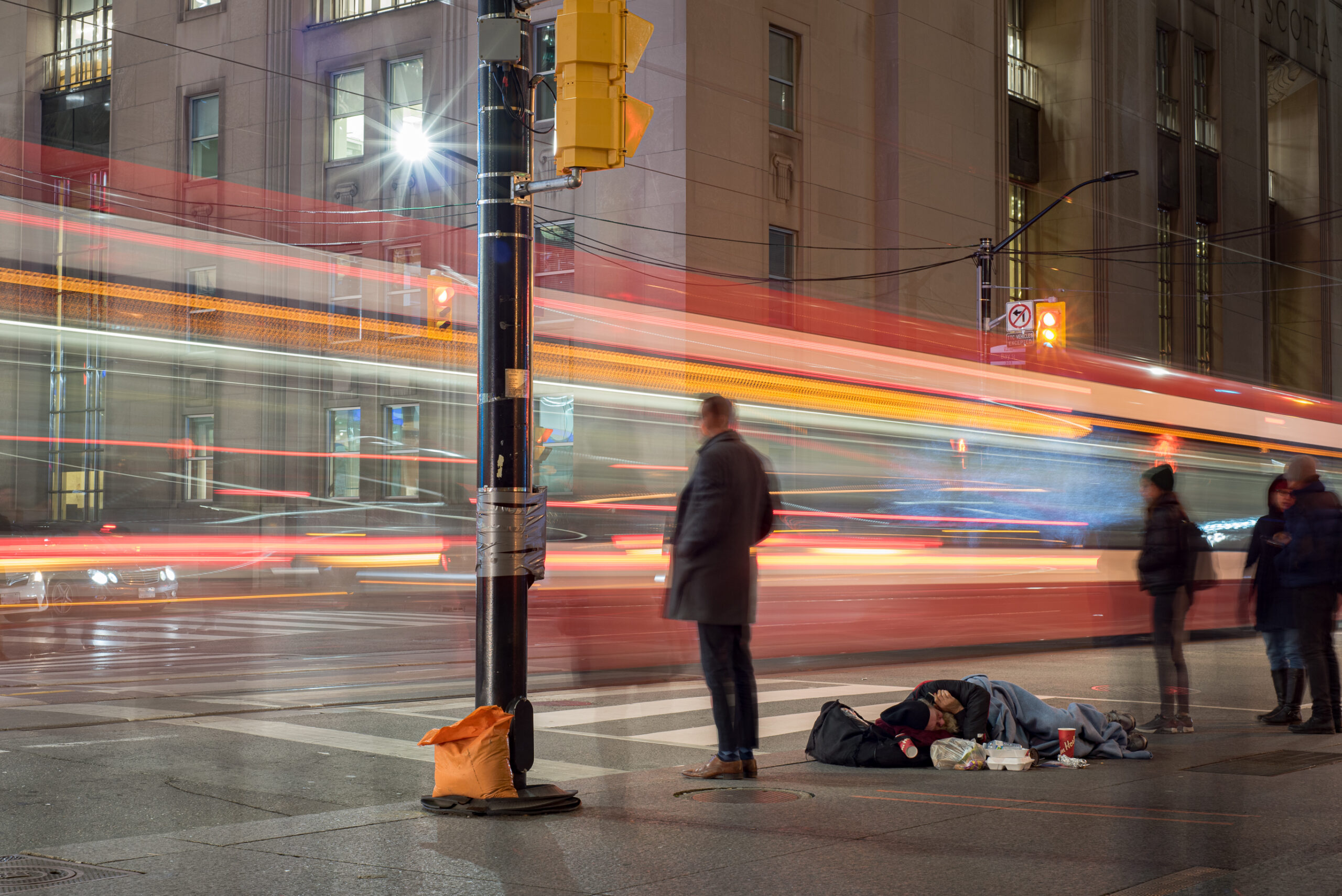 A homeless man lying on a Toronto street in a sleeping bag while a street care passes by.