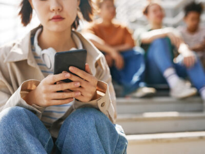 Closeup of teenage girl holding smartphone outdoors while sitting on metal stairs with group of friends in background, copy space