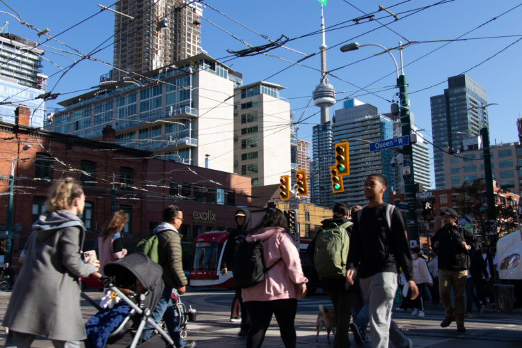 People walk across Queen St. at Spadina Ave. during the afternoon rush hour in downtown Toronto, the CN Tower can be seen in the far background.