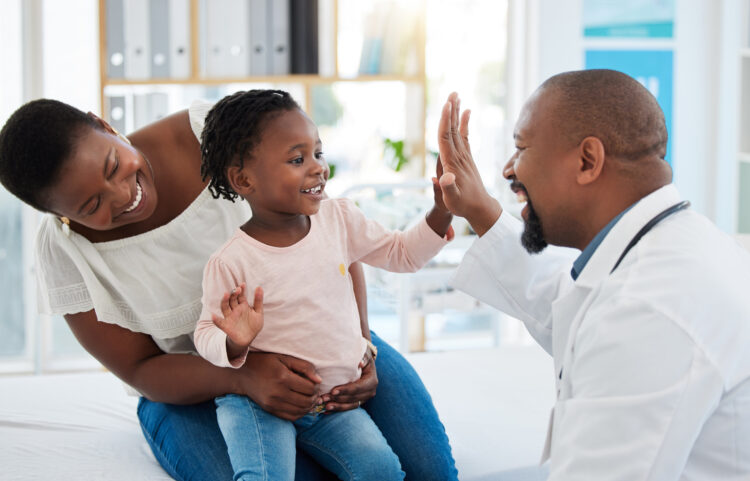 A Black mother smiles while her happy daughter high-fives her doctor in his office.